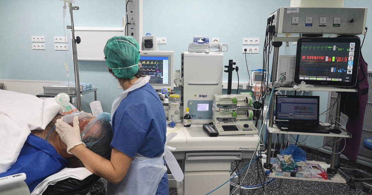 An anesthetist holds a mask over a patient's face at the start of anesthesia, with monitors and the anesthesia machine in an operating room